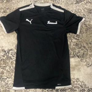 Svart Puma fotbollströja (domare) - Säljer en svart fotbollströja från Puma med vita detaljer och tryck från Svenska Fotbollförbundet på bröstet. Tröjan har texten 'Fotboll ska vara kul!' och 'Landrins Bil' på ryggen. Tillverkad i lätt och ventilerande syntetmaterial, perfekt för träning.