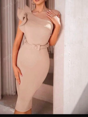 Beige oneshoulder klänning med volang - Säljer en beige, figurnära oneshoulder klänning med snygg volangdetalj över axeln och matchande bälte i midjan. Klänningen har en slits bak och stängs med dragkedja. Perfekt för dig som vill ha en stilren och trendig look.