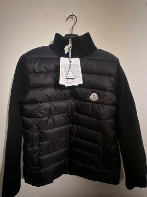 Svart pufferjacka från Moncler - Säljer en svart pufferjacka från Moncler med ribbstickad krage och ärmar. Jackan har quiltad front, dragkedja och två fickor framtill. Klassisk Moncler-logga på bröstet och snygga detaljer. Perfekt för dig som vill ha en stilren och varm jacka.