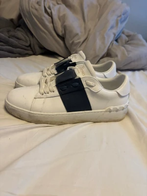 Valentino Garavani vita sneakers - Säljer ett par vita Valentinos Garavani sneakers med bred svart rem över snörningen och ikoniska nitar runt hälen. Skorna har rund tå, platt sula och är tillverkade i skinn. Guldtext på plösen och klassisk streetstil. Perfekta för dig som gillar exklusiva detaljer.