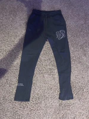 Svarta mjukisbyxor med broderi - Mörk gröna mjukisbyxor med snörning i midjan och broderad logga på ena benet. Textdetalj vid benslutet med 'born in Brooklyn raised on the Streets'. Slimmad passform och mjukt material, perfekta för chill eller streetwear.