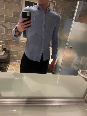 Blå linneskjorta från Polo Ralph Lauren - Säljer en ljusblå slim fit skjorta från Polo Ralph Lauren i linne. Skjortan har klassisk krage, långa ärmar och vita knappar framtill. Ikoniska broderade loggan på bröstet. Perfekt för en clean och stilren look.