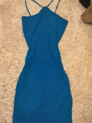 Blå ribbad klänning med smala axelband - Snygg blå ribbad klänning med smala axelband och figurnära passform. Klänningen är kort och har en omlott-detalj vid nederdelen. Perfekt för dig som gillar stilrena och moderna plagg med lite edge.
