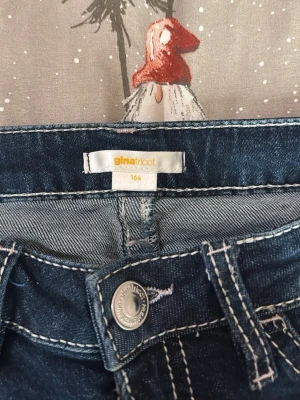 Blå bootcut jeans Gina Tricot 164 - Mörkblå jeans från Gina Tricot med vita kontrastsömmar och snygga detaljer på bakfickorna. Modellen har bootcut-ben och klassisk femficksdesign. Perfekta för dig som gillar en retrovibe och vill ha något som sticker ut lite extra.