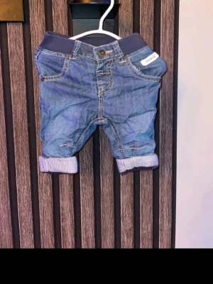 Blå jeansbyxor med mudd från newbie strl 62 - Säljer ett par blå jeansbyxor från Newbie med mjuk mörkblå mudd i midjan och uppvikta ben med randigt foder. Klassisk femficksmodell och knapp framtill. Perfekta för en chill och bekväm stil. Storlek 62