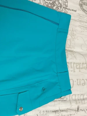 Turkosa golfshorts Calvin Klein - Snygga turkosa golfshorts från Calvin Klein med diskret CK-logga, dragkedja och knapp framtill. Shortsen har bälteshällor och bakficka, tillverkade i ett lätt och stretchigt syntetmaterial som passar perfekt för sommardagar på golfbanan.