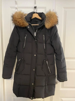 Svart dunjacka Rockandblue med pälskrage - Säljer en svart lång dunjacka från Rockandblue med stor fluffig pälskrage i brunt. Jackan har flera dragkedjor, hög krage och fickor framtill. Perfekt för kalla dagar och har en snygg badge på ärmen. Jackan är quiltad och har långa ärmar. 3799kr nypris 
