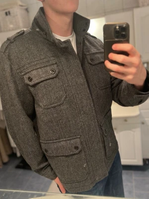 Grå peacoat med fickor och knappar - Säljer en grå rock från Selected Homme i ullblandning med snygg fiskbensmönster. Jackan har fyra stora fickor med lock och knappar, hög krage och axelklaffar. Perfekt för dig som vill ha en stilren och klassisk look till höst och vinter.