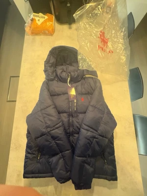 Mörkblå pufferjacka från Polo Ralph Lauren - Säljer en mörkblå pufferjacka från Polo Ralph Lauren med huva och dragkedja framtill. Jackan har det klassiska röda Polo-logotypen broderad på bröstet och två sidofickor med dragkedja. Perfekt för kalla dagar och riktigt snygg till streetwear.