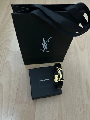 YSL svart läderarmband med guldspänne - Säljer ett stilrent svart armband från Saint Laurent med ikoniskt YSL-logo i guld framtill. Armbandet är gjort i slätt läder och har ett exklusivt guldfärgat spänne där det står 'Saint Laurent Paris'. Perfekt accessoar för dig som gillar lyxiga detaljer.