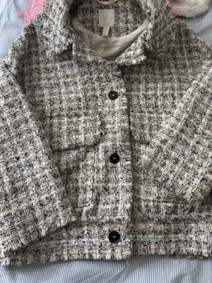 Rutig overshirt i ullmix från H&M - Säljer en snygg rutig overshirt från H&M i en mix av grå, beige och svart. Jackan har stora knappar framtill, krage och en boxig passform. Materialet är tjockt och strukturerat, vilket ger en trendig och mysig känsla. Perfekt att slänga över en hoodie eller t-shirt.