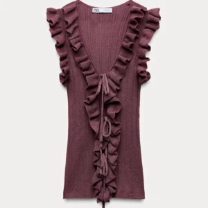 Plommonlila volangtopp från Zara - Supersöt plommonlila topp från Zara med djup v-ringning, volanger längs ringningen och framtill samt knytband. Ribbad struktur och ärmlös design ger en trendig och feminin look. Perfekt att styla med jeans eller kjol för en chic vibe.