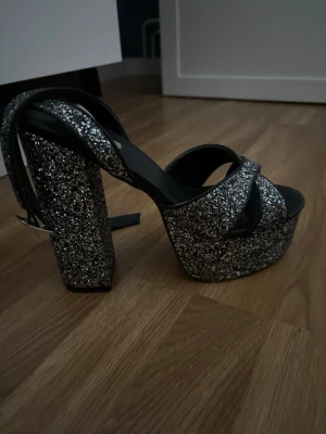 Glittriga platåsandaler från NLY Shoes - Säljer ett par svarta sandaler från NLY Shoes med  platåsula och hög, bred klack. Skorna är täckta av silverglitter som ger en  festlig look. Remmarna korsas över foten och har spänne vid ankeln. Storlek 40. Använt 2 ggr