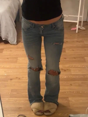 Ljusblå bootcut jeans med slitningar - Säljer ett par ljusblå jeans med bootcut-modell och snygga slitningar vid knäna och låren. Jeansen har klassisk femficksdesign och låg midja. Perfekta för dig som gillar en avslappnad och trendig stil.