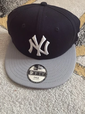 Svart och grå New Era Yankees keps - Säljer en klassisk New Era 9FIFTY snapback med New York Yankees-logga framtill. Kepsen är svart med grå skärm och har justerbar snapback baktill. Perfekt för dig som vill ha en sportig och stilren look.
