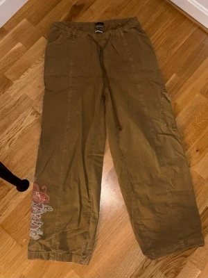 Beiga cargopants med drakbroderi - Unika cargopants från Reclaimed Vintage i beige bomull med dragsko i midjan och coolt broderi av en röd drake på ena benet. Byxorna har sidoficka och relaxed fit. Streetwear och sköna.