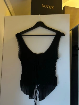 Svart snörad korsettopp med spets - Svart topp i corset-/lingeriestil med spetsdetaljer. Djup urringning och snörning framtill som kan justeras för önskad passform. Breda axelband med spetskant. Figurnära modell som sitter snyggt och ger en feminin silhuett. Perfekt till fest, utekväll eller stylad med jeans/kjol för en trendig look.
