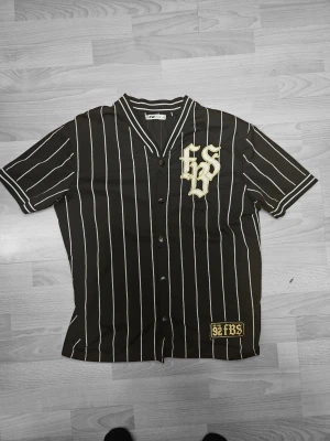 Vintage 90-tals Pinstripe Baseballtröja Svart Vit FBS 92 Oversized - Snygg vintage/90-tals inspirerad pinstripe baseball-shirt i svart med vita ränder 🔥 Guld-broderat "FBS"-logo på bröstet + "EST. 92 FBS"-lapp nederst – perfekt streetwear-känsla! Korta ärmar, knapprad fram, klassisk krage med kontrastsömmar. Oversized/luftig passform – grym att layera eller som statement-plagg. Skick: Väldigt bra begagnat skick – inga hål, inga fläckar, minimalt slitage (lite pyttesmå noppor normalt för åldern). Nyligen tvättad & redo att bäras. Storlek: S (men sitter oversized –