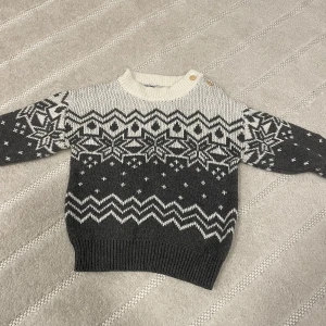Gråvit stickad tröja med mönster - Säljer en grå och vit stickad tröja med snyggt vintermönster och ribbade muddar. Tröjan har långärmade ärmar och rund hals med två knappar på axeln. Perfekt för kalla dagar och riktigt mysig stil. För barn i storleken 74-80