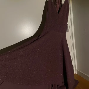 Glittrig vinröd långklänning med spets - Nude Rosa långklänning med glittriga detaljer och djup v-ringning. Klänningen har spets längs sidorna och en elegant, svepande kjol som ger en riktigt lyxig känsla. Perfekt för bal eller fest när du vill sticka ut och känna dig extra glam.