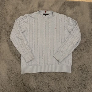 Ljusblå kabelstickad tröja Tommy Hilfiger - Kabelstickad ljusblå tröja från Tommy Hilfiger med rund halsringning och diskret logga på bröstet. Tröjan har ribbade muddar och nederkant, samt klassisk passform och långa ärmar. Perfekt för en stilren och chill look.