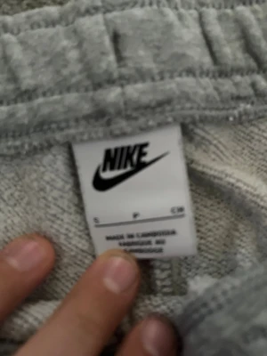 Grå mjukisbyxor från Nike, storlek S - Grå mjukisbyxor från Nike med klassisk logga på vänster lår. Byxorna har snörning i midjan och muddar vid bensluten för en skön passform. Perfekta för chill eller träning, tillverkade i mjukt bomullsmaterial.