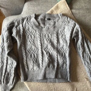 Grå stickad tröja från SHEIN - Grå stickad tröja från SHEIN med snyggt kabelmönster och ribbade muddar vid hals, ärmslut och nederkant. Modellen har rund hals och långa ärmar, perfekt för att hålla dig varm och stilren under kyliga dagar.