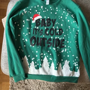 Grön jultröja med tryck, storlek M - Rolig grön jultröja med texten 'Baby it's cold outside' och snöflingor samt vita granar längst ner. Trycket har även en liten tomteluva. Tröjan är långärmad och tillverkad i 100% polyester, perfekt för att maxa julstämningen. Lite noppig på armarna 
