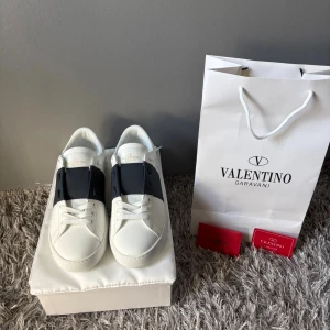 Valentino Garavani vita sneakers - Snygga vita sneakers från Valentino Garavani med svart detalj över snörningen. Skorna har rund tå, platt sula och är tillverkade i skinn. Klassisk och stilren design med logotyp på tungan och undersidan. Levereras med originalpåse och certifikat. Pris kan diskuteras…