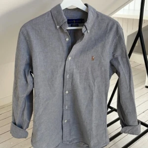 Grå slim fit skjorta från Ralph Lauren - Snygg grå skjorta från Ralph Lauren i slim fit-modell, tillverkad i stretchig bomull. Klassisk oxfordkrage, knappar framtill och ikonisk broderad logga på bröstet. Perfekt för dig som gillar stilrena och tidlösa plagg.