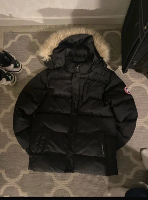 Svart pufferjacka från Canada Goose - Svart pufferjacka från Canada Goose med fluffig huva och beige pälsdetalj. Jackan har dragkedja, knappar och två stora fickor framtill. Klassisk patch på ärmen och snygg, oversized look. Perfekt för kalla vinterdagar och riktigt varm.