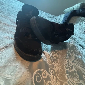 Svarta Nike Jordan 4 sneakers - Säljer ett par svarta Nike Air Jordan 4 sneakers med klassisk Jumpman-logga på plösen och yttersulan. Skorna har snörning, meshpaneler på sidorna och en robust platt sula. Materialet är en mix av mocka och syntet, vilket ger en cool och sportig vibe.