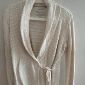 Crèmefärgad stickad omlottkofta från Bondelid - Mysig beige stickad omlottkofta från Bondelid med ribbad struktur och knytband i midjan. Koftan har lång ärm och en bred krage som ger en chill och snygg look. Perfekt att slänga på sig när det är lite kyligt ute