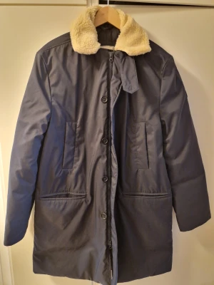 Mörkblå parkajacka från Boomerang Large - Parkajacka från Boomerang i mörkblått med mjuk teddykrage i beige. Jackan har både dragkedja och knappar framtill, två stora fickor och en rak, längre passform. Perfekt för kalla dagar med sin fodrade insida och stilrena look.