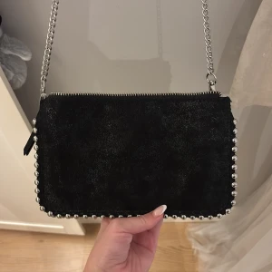 Svart axelväska med silverkedja - Snygg svart axelväska i mocka med silvriga nitar runt kanterna och en silverfärgad kedja som axelrem. Väskan har dragkedja upptill och en rektangulär form som💗