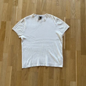 Vit ribbad t-shirt från H&M - Vit t-shirt från H&M i regular fit med ribbad struktur och rund hals. Tillverkad i en mjuk mix av 50% bomull och 50% viskos. Klassisk kortärmad modell med diskreta vertikala ränder som ger en clean och fräsch look.