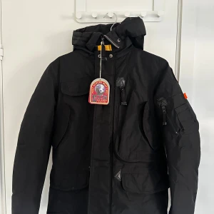 Svart parkajacka från Parajumpers - Svart parkajacka från Parajumpers med huva, flera fickor och coolt PJS-märke på ärmen. Jackan har dragkedja och knappar framtill, samt orange detalj på ärmen. Perfekt för kalla dagar med sin rejäla och funktionella design. Pris kan diskuteras. Kvitto finns