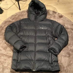 Svart pufferjacka från Sail Racing - Svart pufferjacka från Sail Racing med huva och diskret logga på bröstet. Jackan har breda, quiltade sektioner och dragkedja framtill. Materialet är syntet och dun, perfekt för kalla dagar. Snygg och enkel design som funkar till det mesta. 2 hål som är fixade med lappar annars skit fräsch.( sitter bra på 186 och väger 73 typ😀