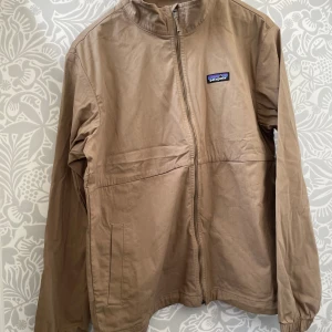Beige vindjacka från Patagonia - Säljer en beige vindjacka från Patagonia i storlek M. Jackan har stilren design med ståkrage, hel dragkedja och två sidofickor. Materialet är slitstark bomull och jackan har ett broderat Patagonia-märke på bröstet. Perfekt för dig som gillar clean och enkel stil. Säljes pga för liten för mig, nypris 2100:- köpt för 1700:- 