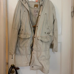 Beige parkajacka från Parajumpers - Säljer en beige parkajacka från Parajumpers med coolt patch på ärmen och flera stora fickor framtill. Jackan har huva, meshfoder och dragkedja. Perfekt för dig som gillar utility-vibe och vill ha något som sticker ut lite extra. Jackan är i L men jag som är i storlek m passar den perfekt så skulle säga den är liten i storleken 