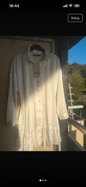 Vit skjortklänning med volang från Zara - Säljer en vit skjortklänning från Zara med klassisk krage, knappar hela vägen framtill och volang nedtill. Klänningen har långa ärmar och är tillverkad i ett glansigt, mjukt material som ger en lyxig känsla. Perfekt för dig som gillar stilrena och trendiga plagg. Storlek M passar dock en S eller xs då man kan spänna den/knyta 