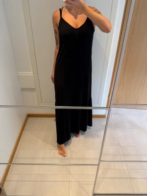 Svart långklänning med korsade band - Svart långklänning med smala axelband som korsas snyggt i ryggen. Klänningen har v-ringad hals och är tillverkad i mjuk polyester med lite stretch för skön passform. Stilren och enkel design som ger en cool vibe. Maxiklänning med smala dubbla korsade axelband och voluminös kjol. Perfekt till t.ex. nyår!🥂