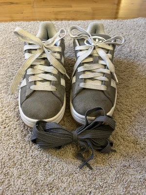 Grå Adidas campus 00 unisex - Säljer ett par grå adidas campus sneakers med klassiska vita ränder på sidorna och vit logga på plösen. Skorna har rund tå, platt sula och snörning. Ovandelen är i mocka och insidan är mjukt vadderad för extra komfort. Perfekt för dig som gillar stilrena och snygga skor. Skorna kommer även med ett par gråa snören. Knappt använda. Säljs på grund av att det var fel storlek för mig. 