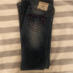 True Religion blå jeans med röd söm - Snygga blå jeans från True Religion med coola röda kontrastsömmar och klassisk logga på bakfickan. Jeansen har silverfärgade knappar och en faded tvätt som ger en avslappnad vibe. Passar dig som gillar detaljer och streetstyle. ALDRIG ANVÄNDA