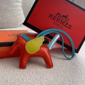  Pegasus bag charm i skinn - Superfin  Pegasus bag charm i form av en häst med vingar. Gjord i mjukt skinn i orange, blått och gult. Perfekt accessoar för att ge din väska en unik och färgglad touch. Kommer med originalbox.