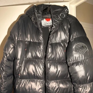 Svart pufferjacka från Zavetti Canada XL - Snygg svart pufferjacka från Zavetti Canada i storlek XL. Jackan har broderad logga på ärmen, huva med broderad text, och flera dragkedjor med coola detaljer. Materialet är glansigt och känns riktigt robust, perfekt för kalla dagar. Insidan har mönstrad lining med logga.