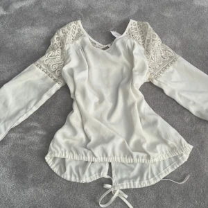 Vit blus med spets och snörning - Säljer en vit blus med långa ärmar och snygga spetsdetaljer på axlar och överarmar. Blusen har rund halsringning och snörning nedtill som ger en cool, justerbar passform. Materialet är lätt och flowy, perfekt för dig som gillar romantiska detaljer.