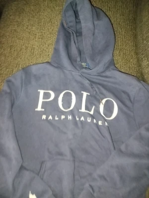 Marinblå hoodie från Polo Ralph Lauren - Snygg marinblå hoodie från Polo Ralph Lauren med stor vit logotyp på bröstet och klassisk polospelare broderad på ärmen. Tröjan har huva med snörning och en känguruficka framtill. I storlek Medium för herr