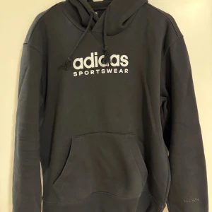 Svart hoodie från Adidas Sportswear - Svart hoodie från Adidas Sportswear med vit logga på bröstet och broderad 'ALL SZN' på ärmen. Tröjan har huva med snörning och en stor magficka. Mjuk insida i bomull och polyester, perfekt för chill eller träning. 