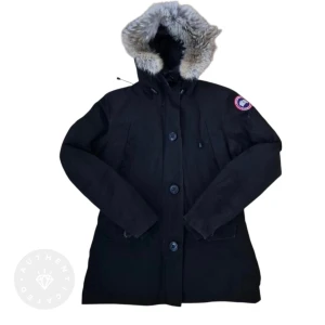 Svart dunparkas från Canada Goose montebello - Svart dunparkas från Canada Goose med päls på huvan och klassisk logga på ärmen. Jackan har långa ärmar, stora knappar framtill och dragkedja under knappslån. Fodrad insida och justerbar midja. Perfekt för kalla vinterdagar.